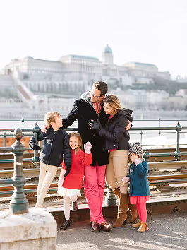 Family of five standing on a riverside promenade with historic buildings in the background, warmly dressed and interacting cheerfully.