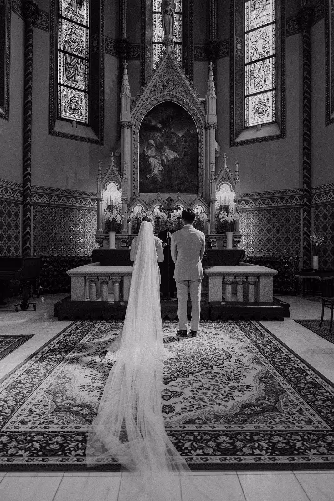Bride and groom standing at the altar in a church with stained glass windows and ornate decorations during a wedding ceremony.