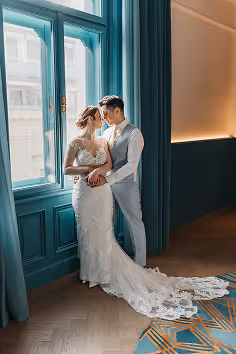 Bride in a white lace wedding gown and groom in a light gray suit standing close by a large window with teal curtains.