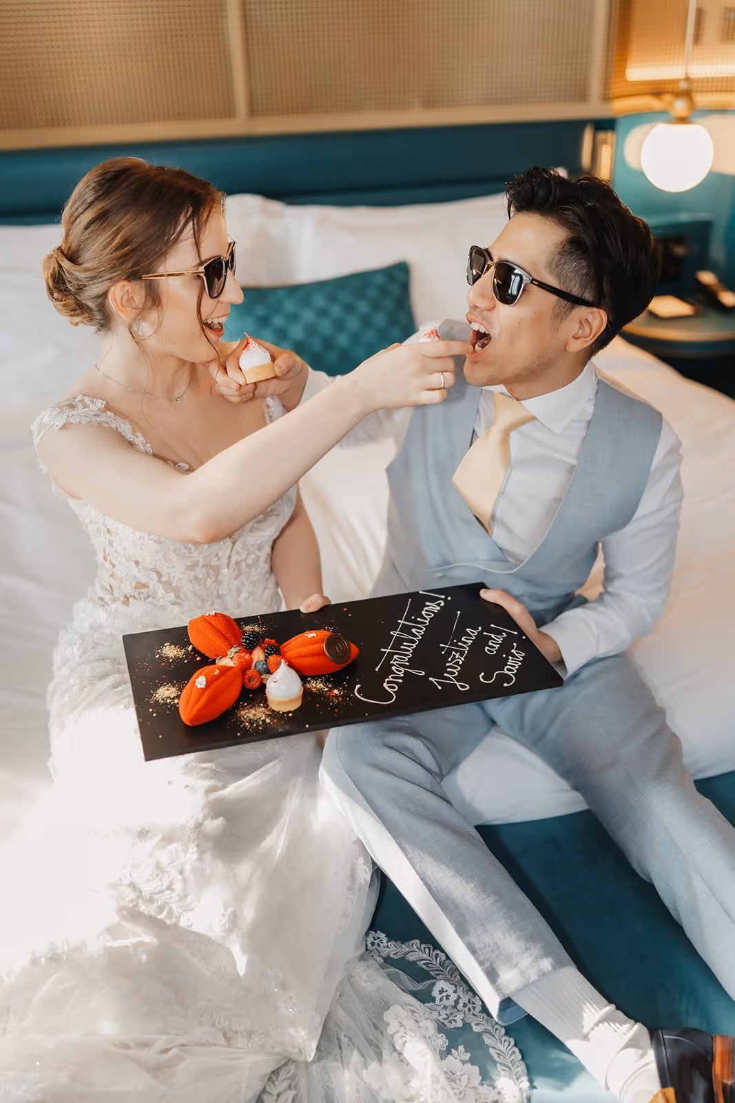 Bride in wedding dress and groom in suit with sunglasses feeding each other small desserts on bed, holding a congratulatory dessert board.