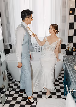 Bride in lace wedding gown playfully tugging groom's tie as they stand in a bathroom with black and white checkered floor.