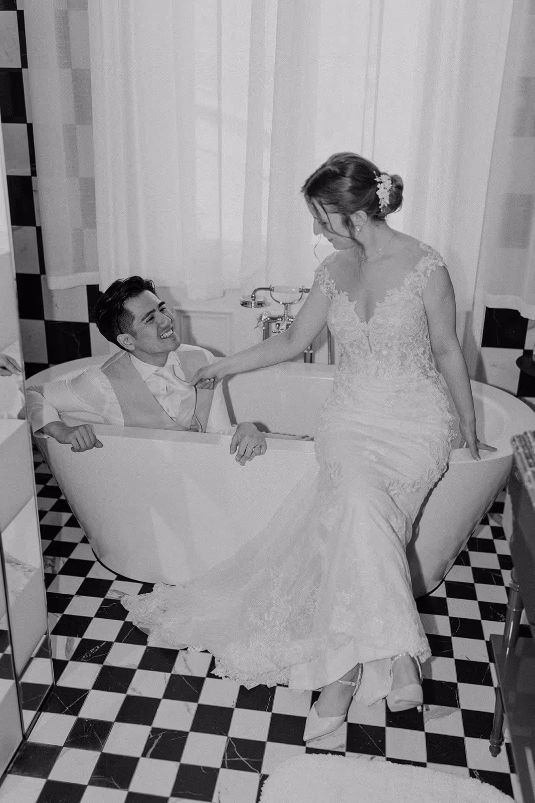 Bride in lace wedding dress sitting on the edge of a bathtub, holding hands and smiling at groom inside the tub on checkered floor.