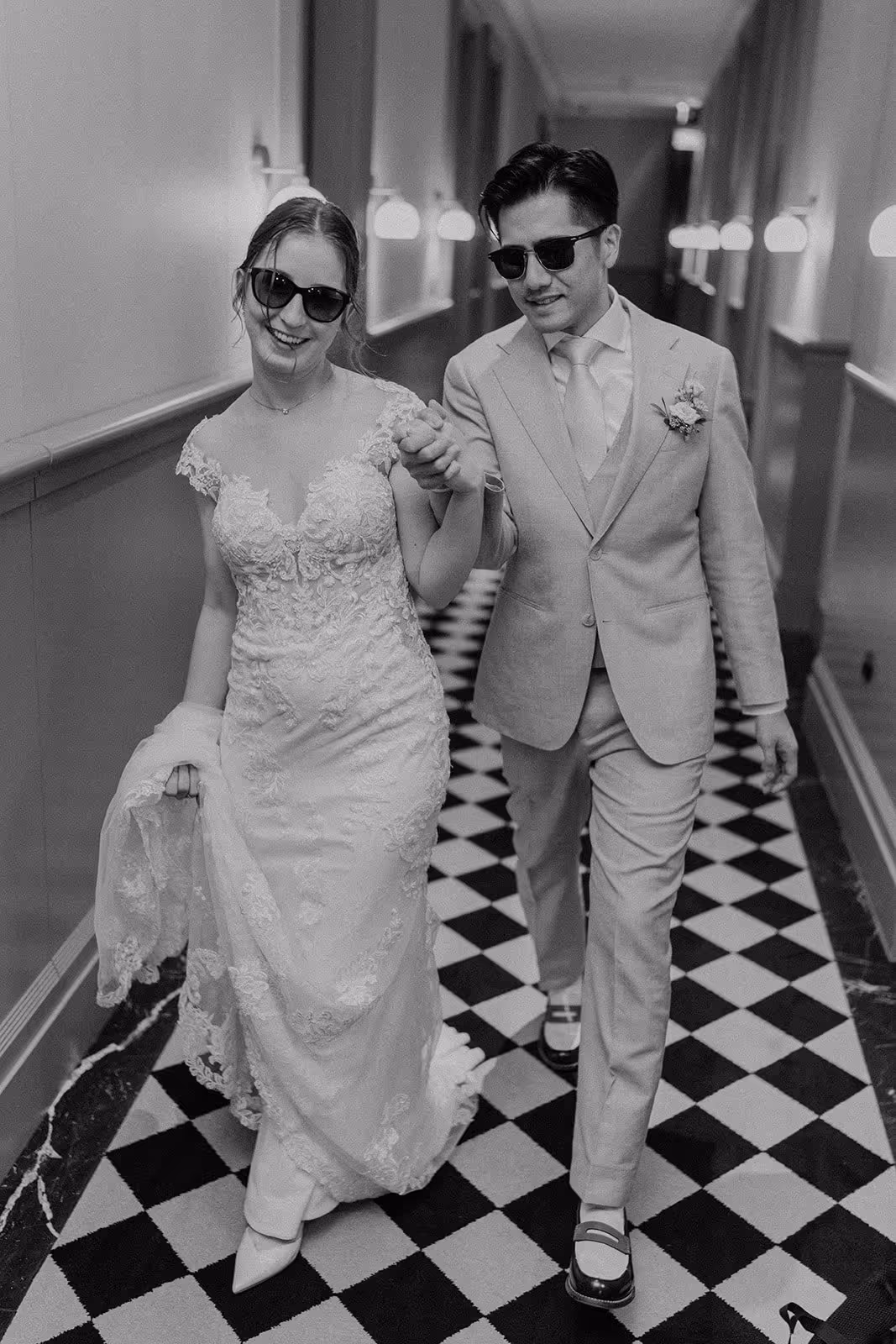 Bride and groom wearing sunglasses, walking hand in hand down a corridor with a checkered floor, bride in lace wedding dress and groom in suit.