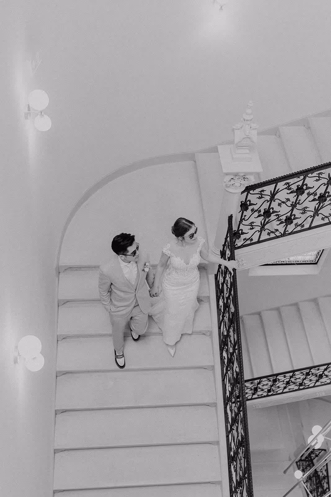 Black-and-white photo of a bride and groom holding hands while walking up a staircase with ornate railing.