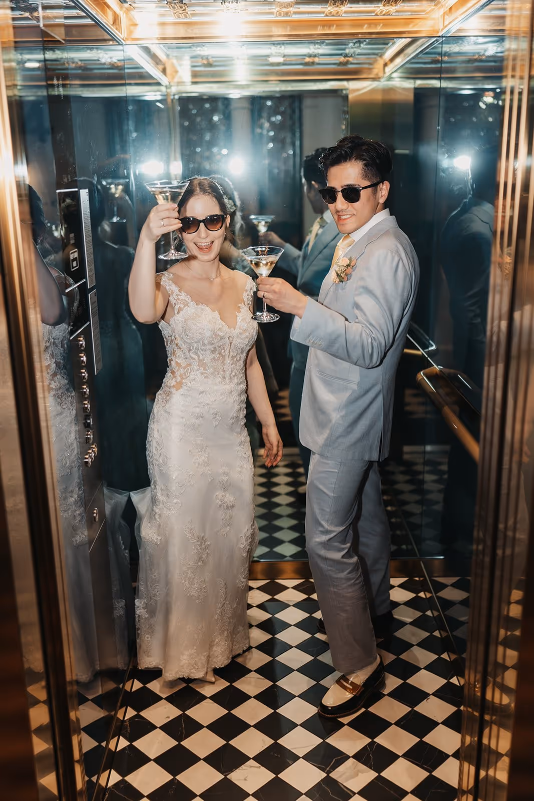 Bride in a lace wedding dress and groom in a light gray suit wearing sunglasses, raising martini glasses in a mirrored elevator with black-and-white checkered floor.
