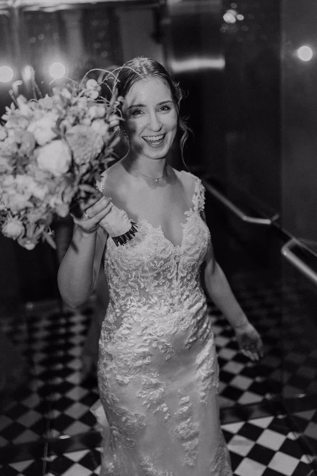 Smiling bride in a lace wedding dress holding a bouquet, standing on a checkered floor.