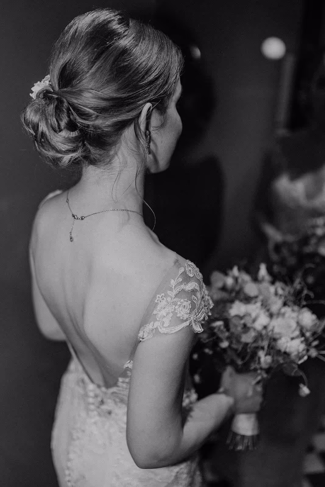 Bride with lace wedding dress holding a bouquet, photographed from behind in black and white.