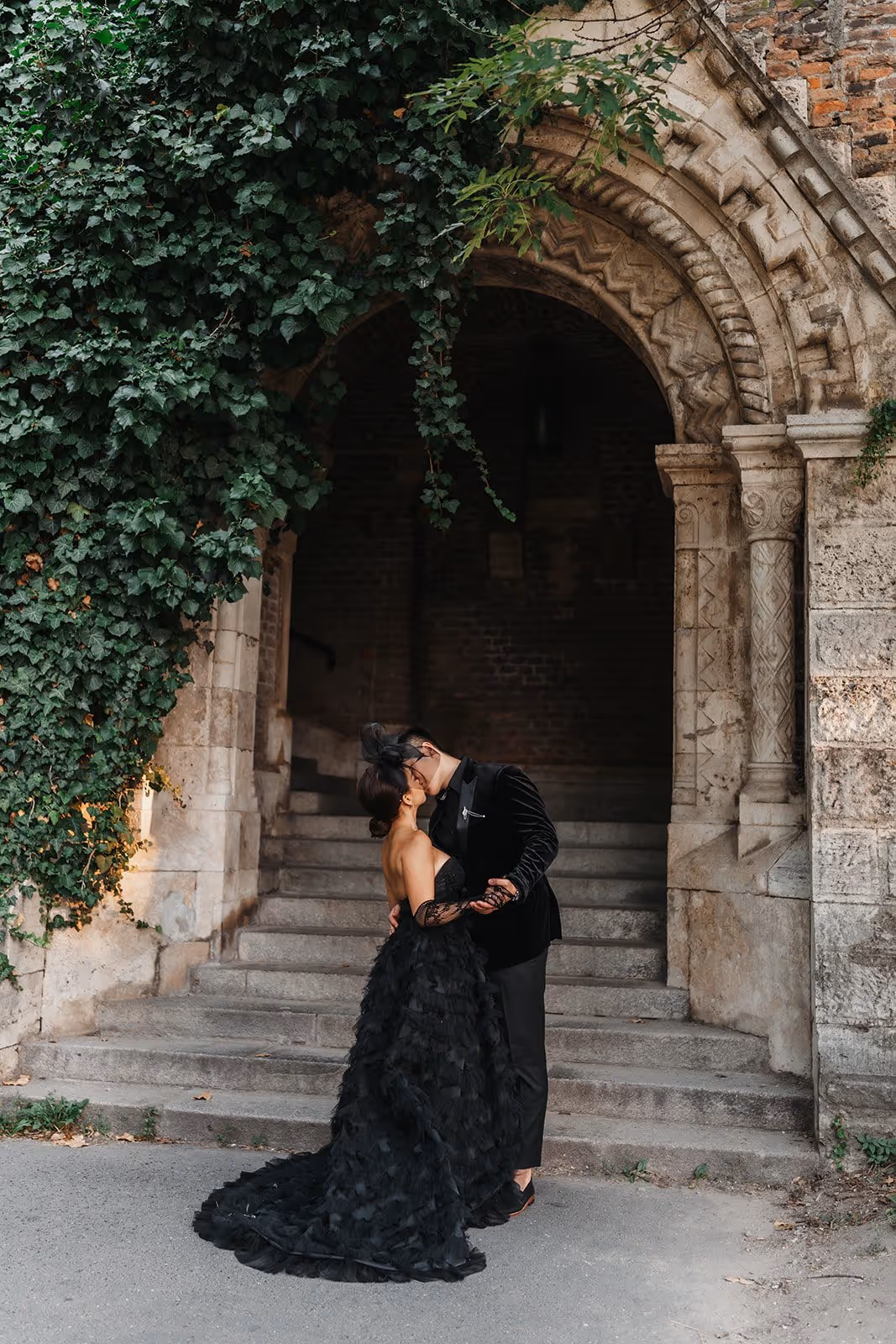 Couple dressed in elegant black attire sharing a kiss in front of stone steps and an archway covered with green ivy.