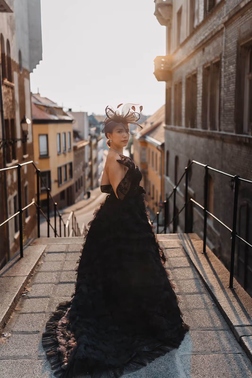 Woman in an elegant black gown and matching hat standing on stone steps in an old city street at sunset.