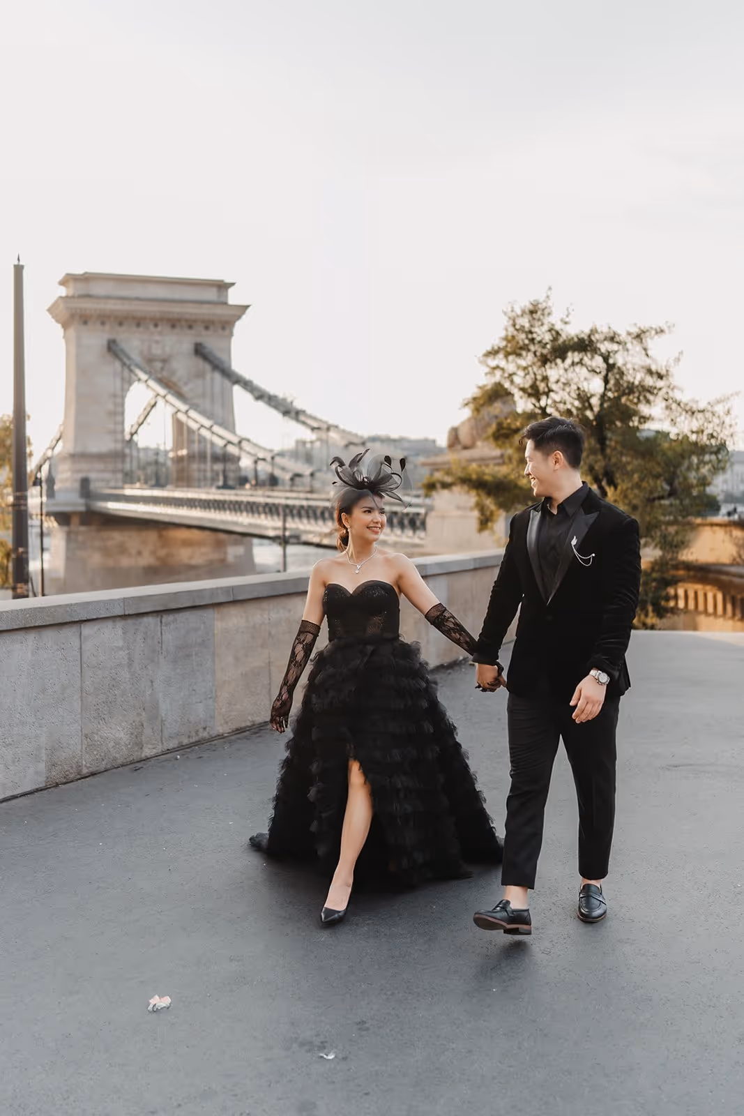 Couple dressed in elegant black attire holding hands and walking on a paved path near a suspension bridge at sunset.