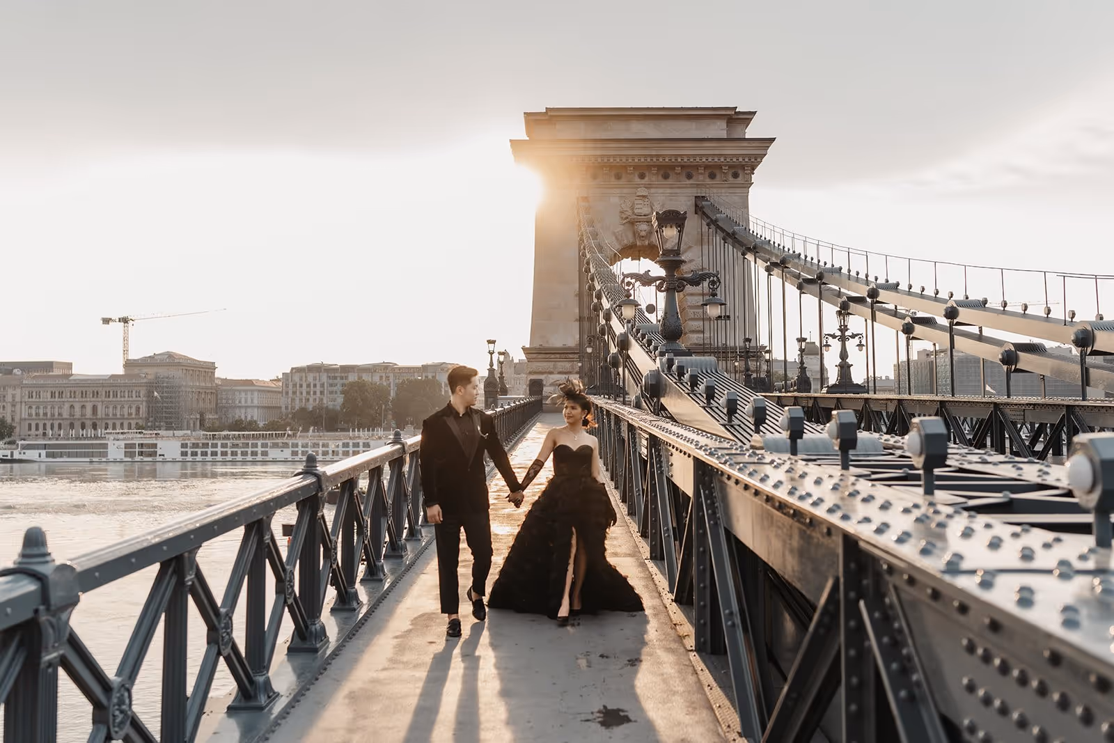 A couple dressed in elegant black attire holding hands and walking on a historic suspension bridge at sunset.