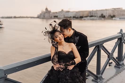 Couple dressed in elegant black attire embracing on a bridge with a river and cityscape in the background at sunset.