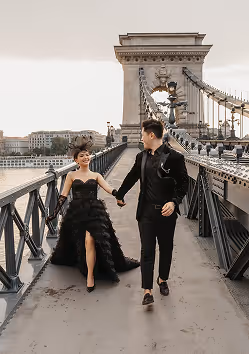 Couple dressed in formal black attire walking hand-in-hand on a bridge with cityscape in the background.