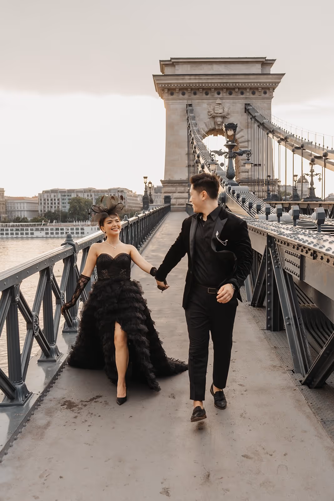 Couple dressed in elegant black attire holding hands and walking on a historic suspension bridge at sunset.