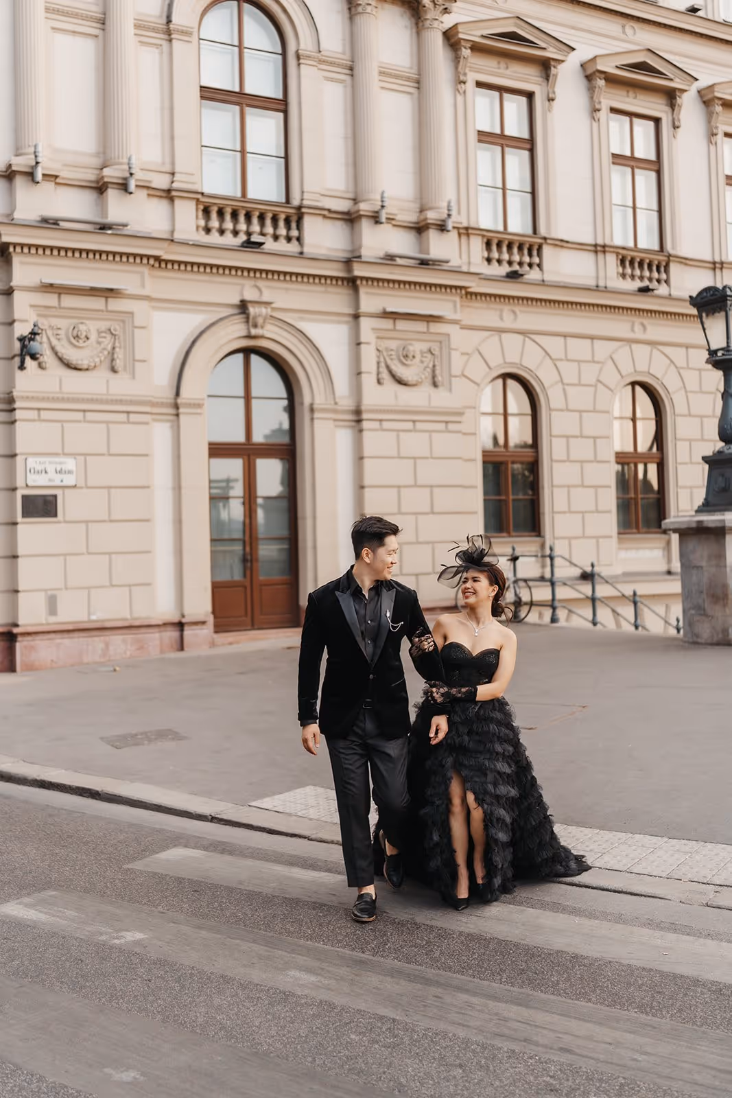 A couple dressed in elegant black evening attire walking arm in arm on a street in front of a historic stone building.
