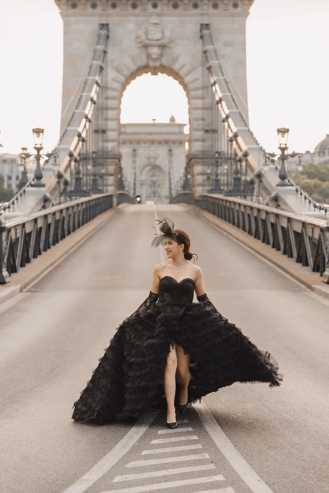 Woman in a flowing black strapless gown with gloves and a feathered fascinator walking on a historic suspension bridge.