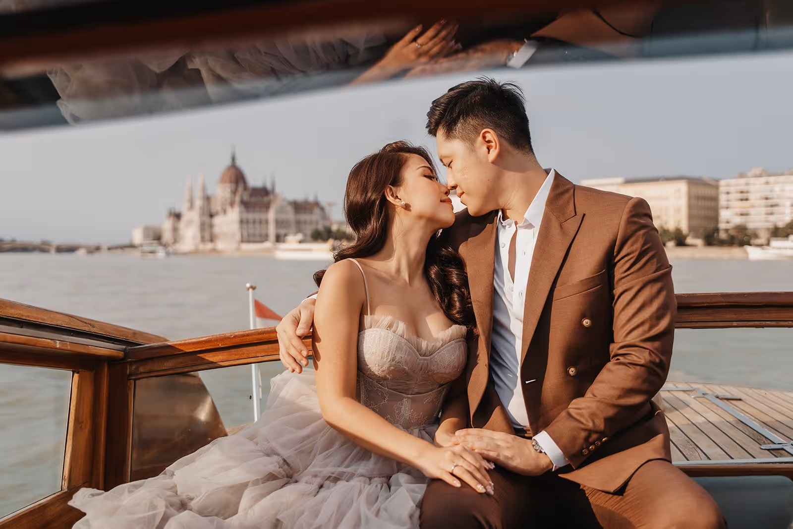Couple in formal attire sitting on a boat, leaning in for a kiss with a historic building across the river in the background.