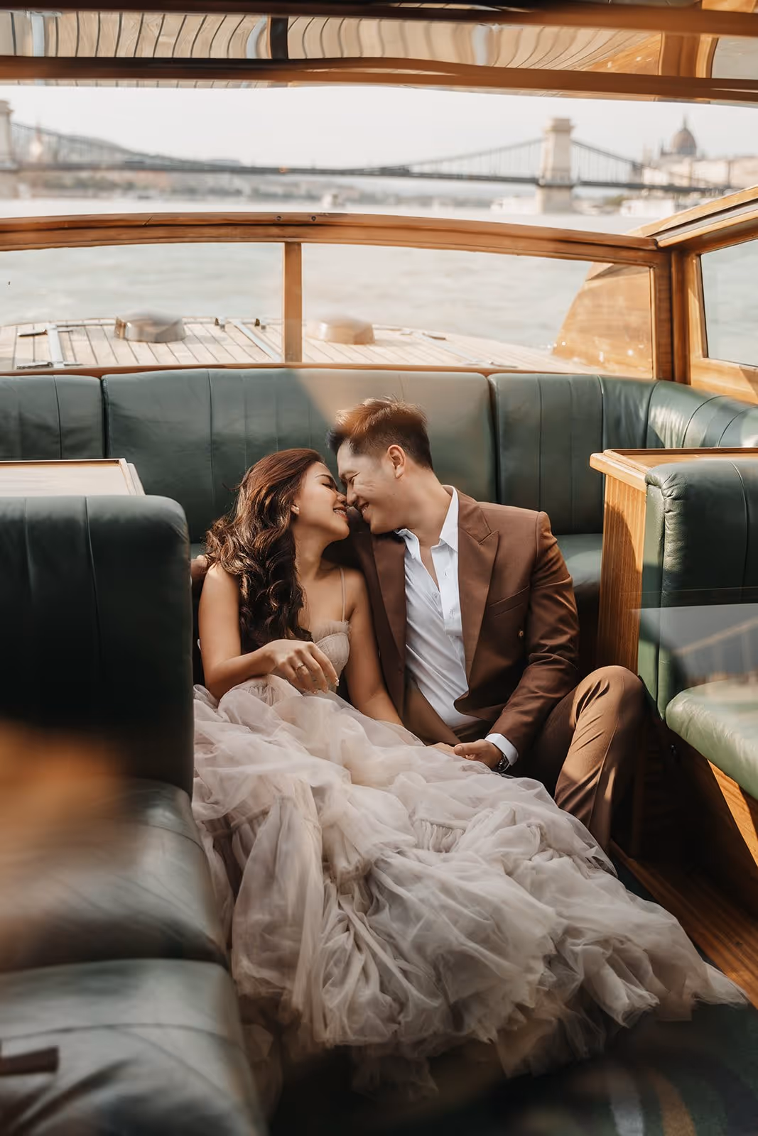 Couple in elegant attire sitting closely on a boat with a river and a suspension bridge in the background.