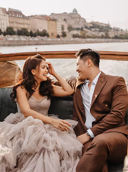 Couple smiling and sitting closely together on a boat with a cityscape and river in the background.
