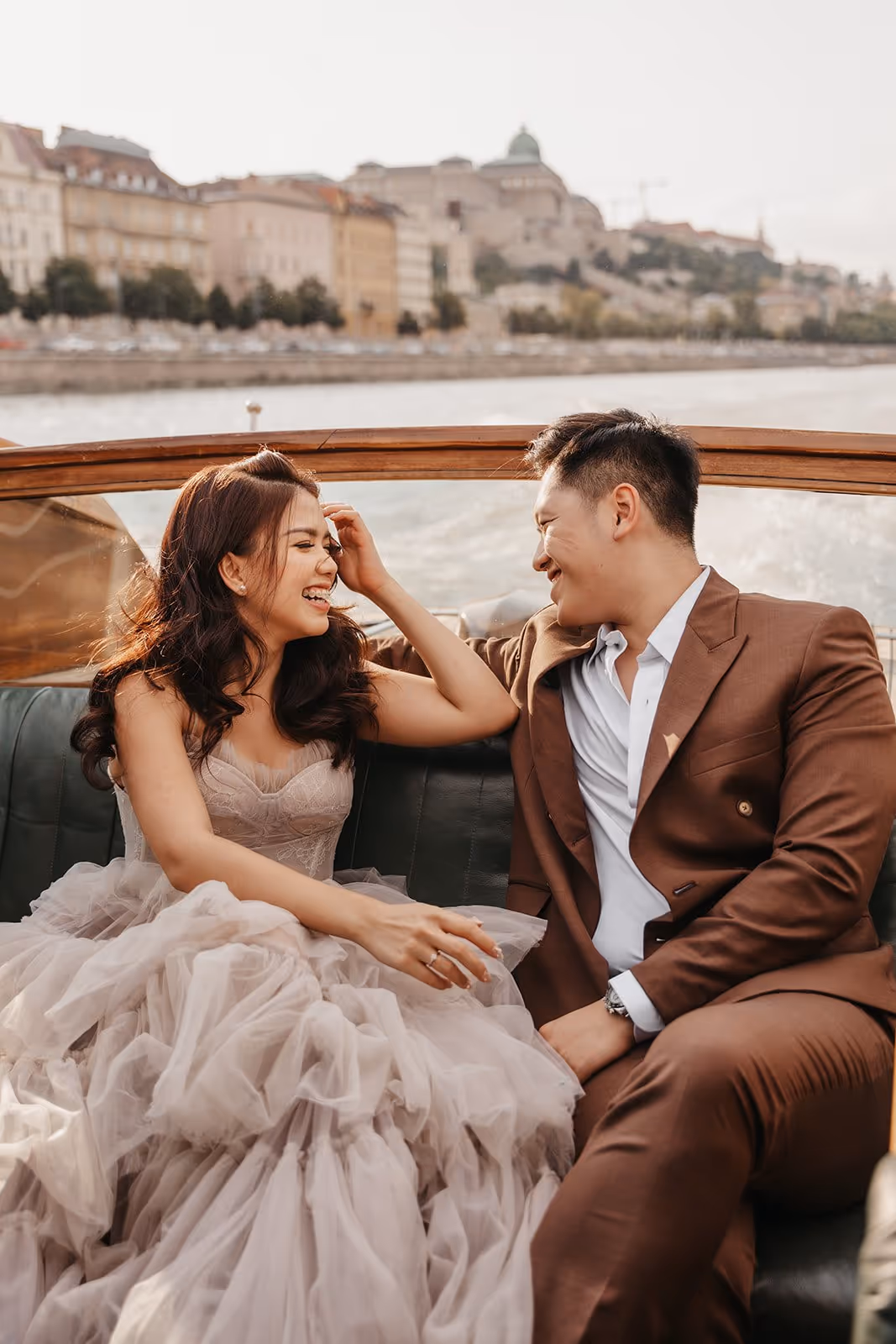 Smiling couple dressed formally and sitting close together on a boat with city buildings and water in the background.