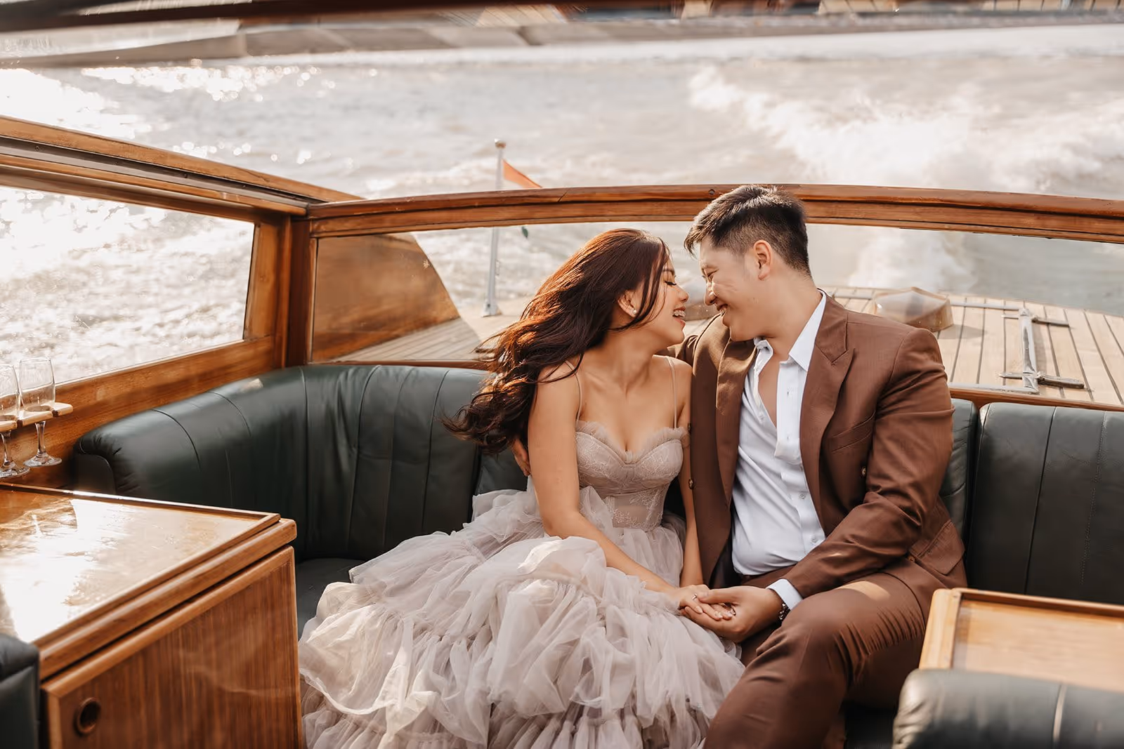 Couple dressed elegantly smiling and holding hands while sitting on a boat with water in the background.