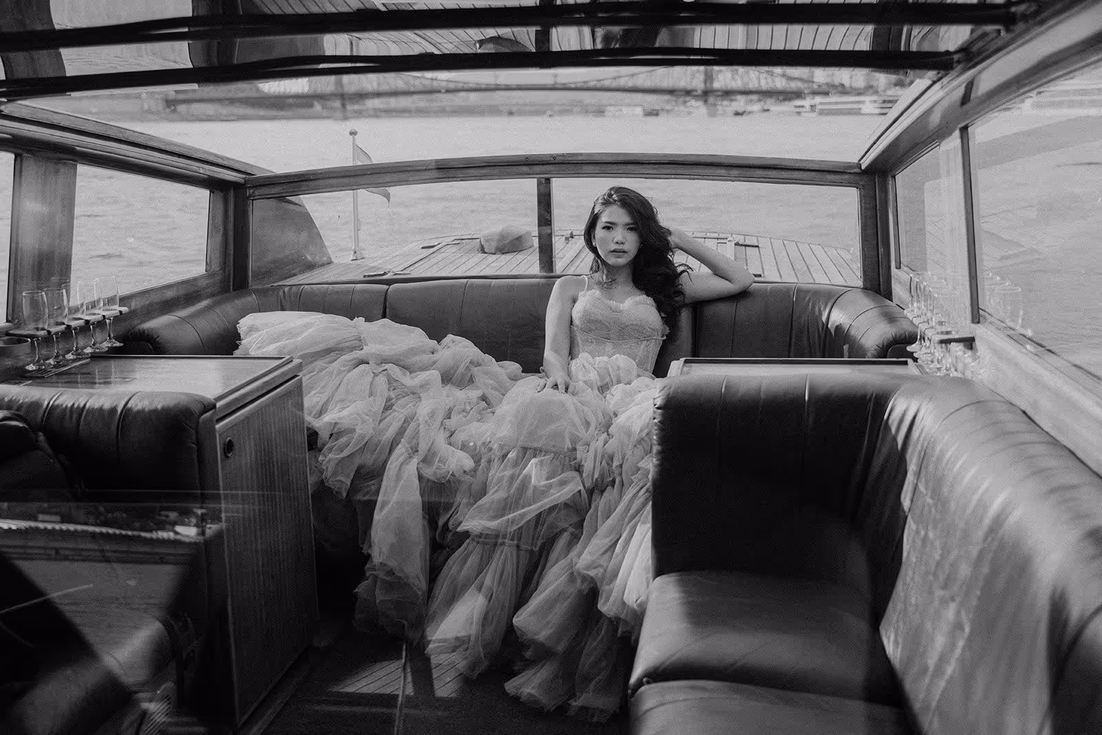 Woman wearing an elaborate dress reclining on a leather bench inside a boat with water visible outside the windows.