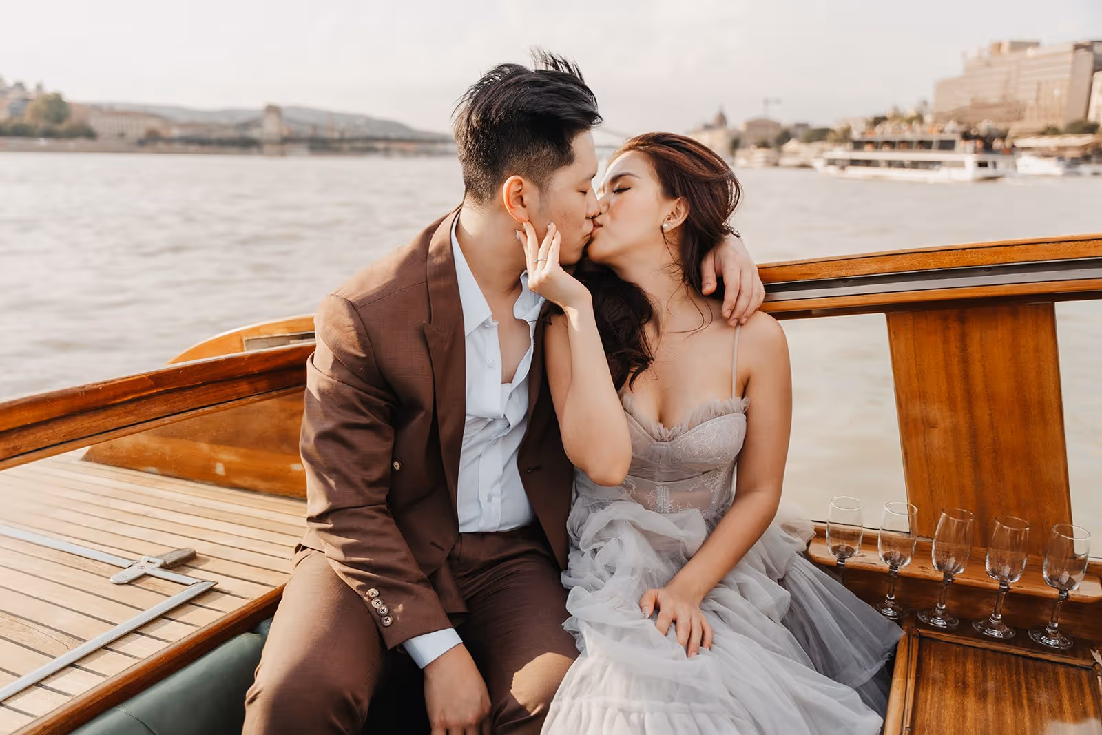 Couple dressed elegantly kissing on a wooden boat with water and cityscape in the background.