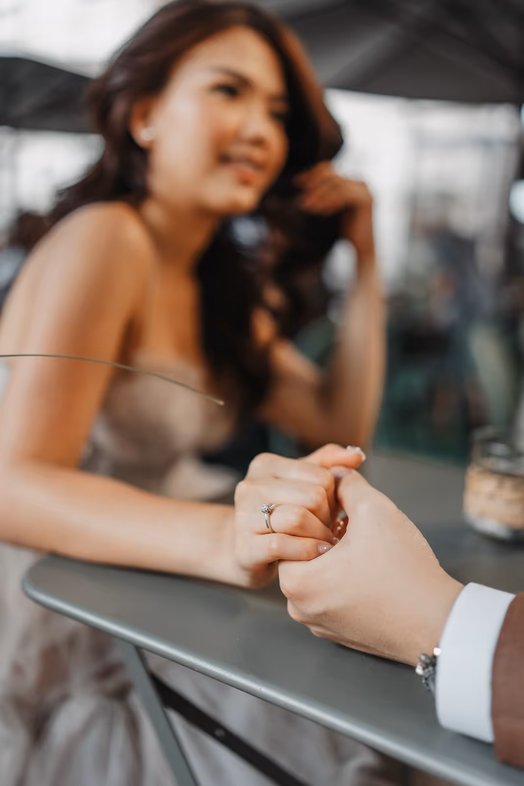 Close-up of a couple holding hands at a table, with a woman wearing an engagement ring and smiling in the background.