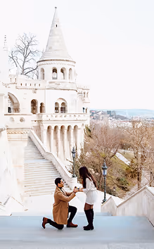 Man kneeling and proposing to woman on stairs in front of a castle-like building with a tower.