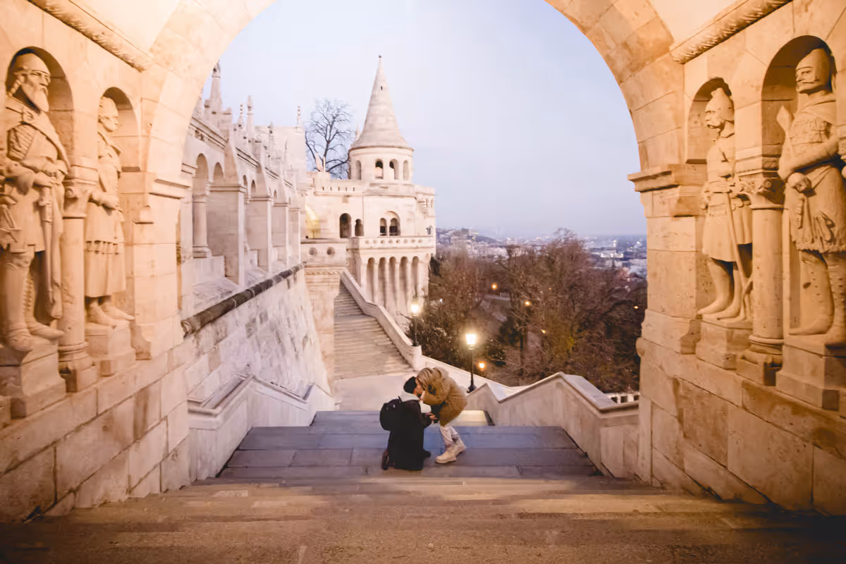 Couple kissing on stone steps framed by archway with carved statues and castle-like architecture in the background.