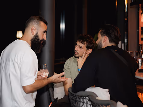 Three men engaged in conversation at a bar, one holding a glass, two sitting on bar stools.