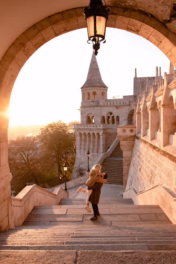 Man lifting woman on stone steps framed by an arch with historic castle-like architecture at sunset.