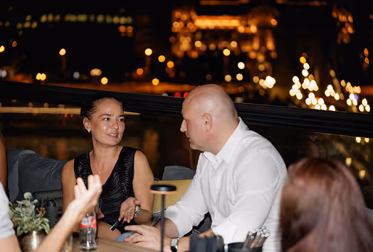 A woman and a man engaged in conversation at an outdoor nighttime setting with blurry city lights in the background.