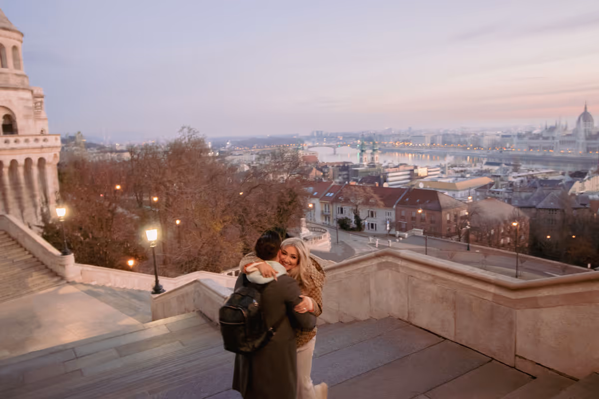 Couple hugging on stone steps overlooking a cityscape with a river and bridge at dusk.