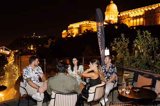 Group of five people sitting and talking on a rooftop terrace at night with a cityscape and illuminated historic building in the background.