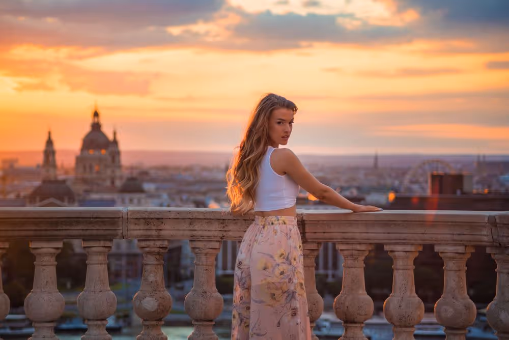 Young woman with long hair wearing a white crop top and floral skirt leaning on a stone balcony railing at sunset overlooking a cityscape.