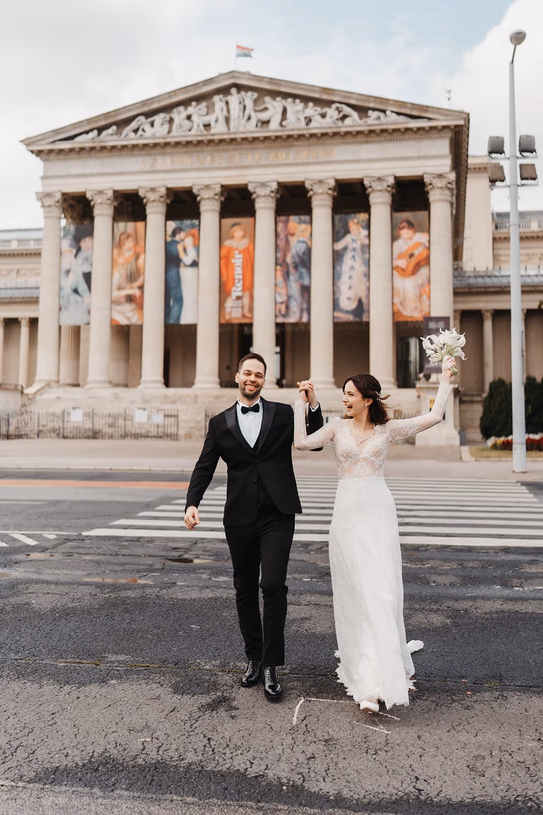 Happy bride and groom holding hands and walking on a crosswalk in front of a grand building with tall columns and art banners.