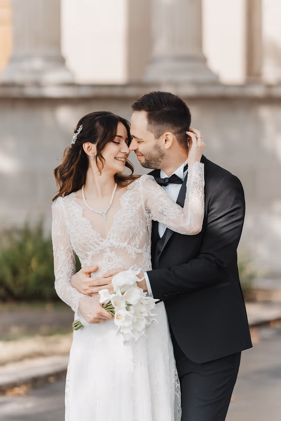 Bride in lace wedding dress holding white calla lilies embraced by groom in black tuxedo, both smiling with foreheads touching.