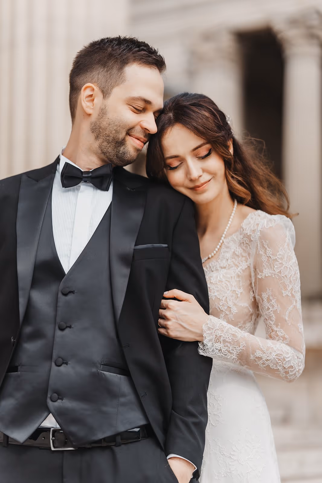 Bride in lace wedding dress gently resting her head on groom's shoulder, who is wearing a black tuxedo and bow tie.