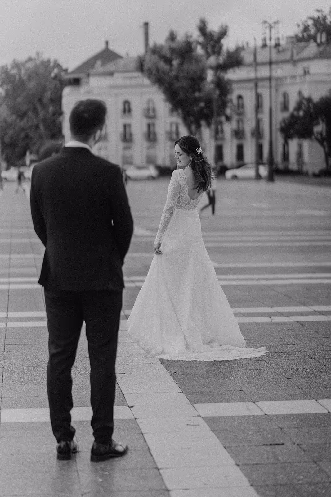 Bride in a lace long-sleeve wedding dress looking back while groom in a dark suit watches her in a city square.