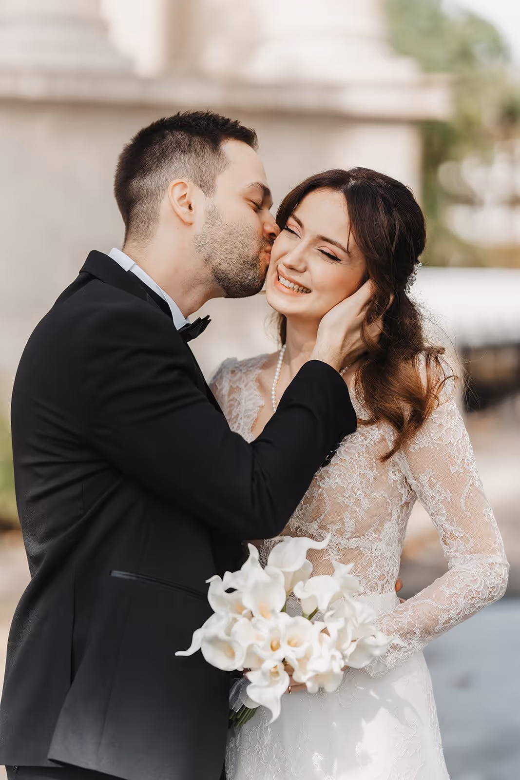 Groom in black tuxedo kissing smiling bride in lace dress holding white flower bouquet.