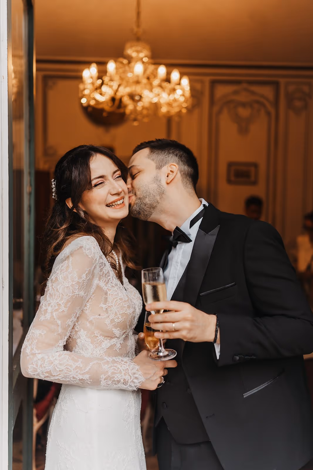 Groom in tuxedo kissing smiling bride in lace wedding dress holding champagne glasses indoors under chandelier.