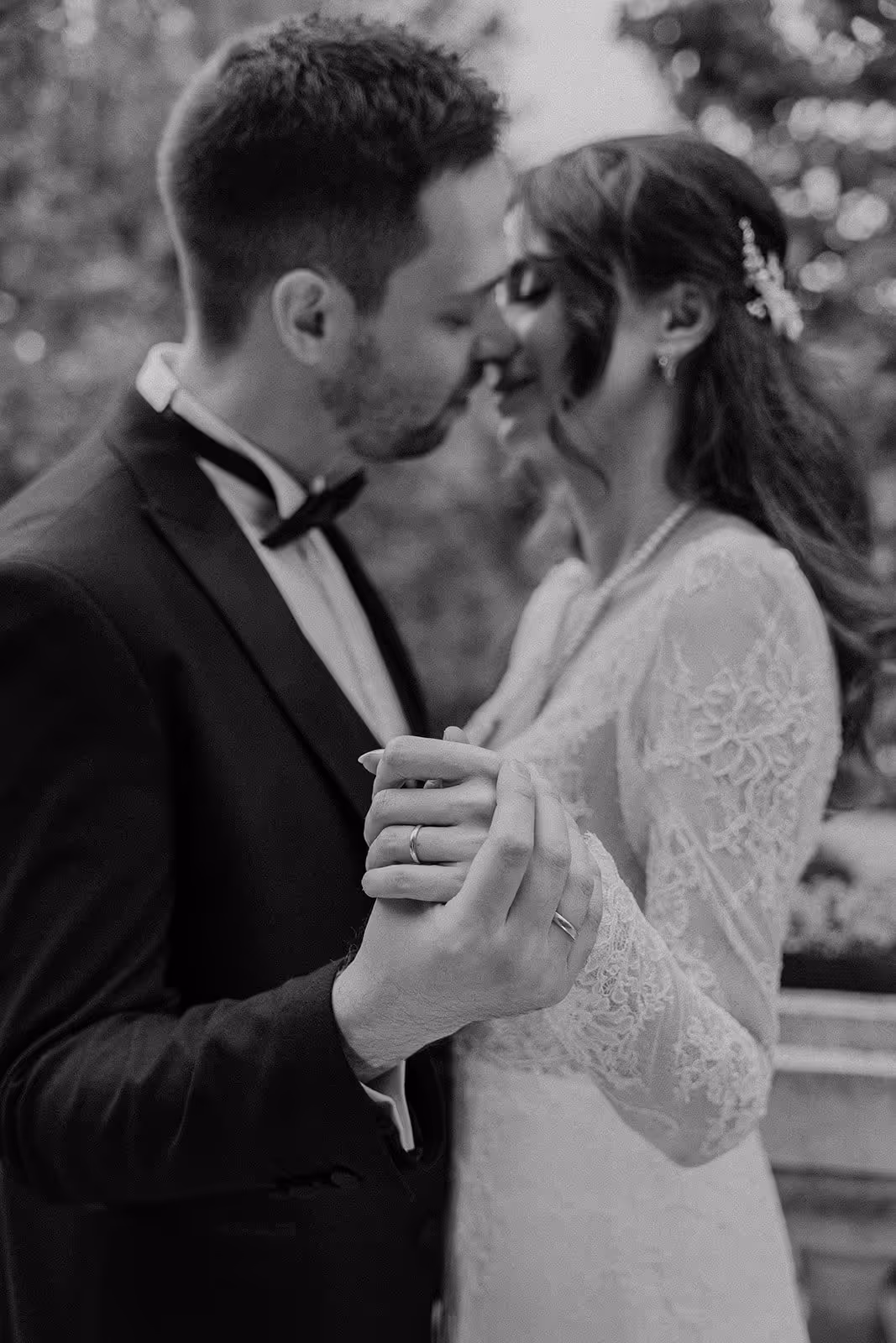 Bride and groom in wedding attire holding hands and leaning in close in a tender moment outdoors.