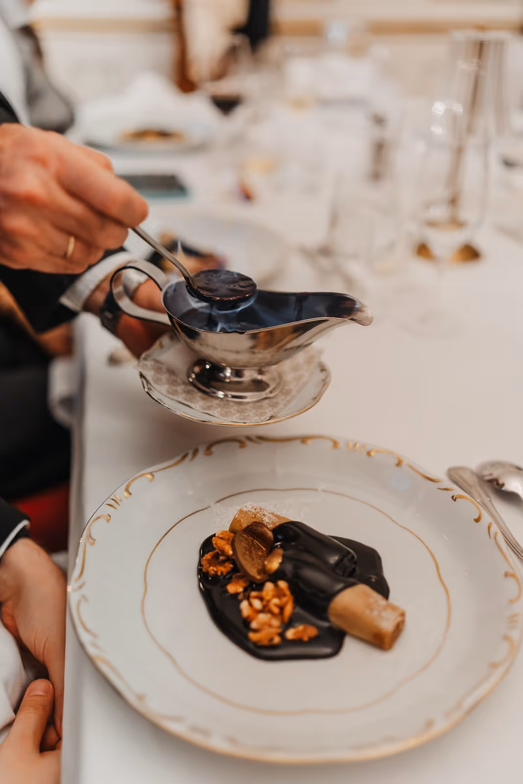 Hand pouring flamed sauce from a metal gravy boat onto plated gourmet dessert with chocolate sauce and nuts.