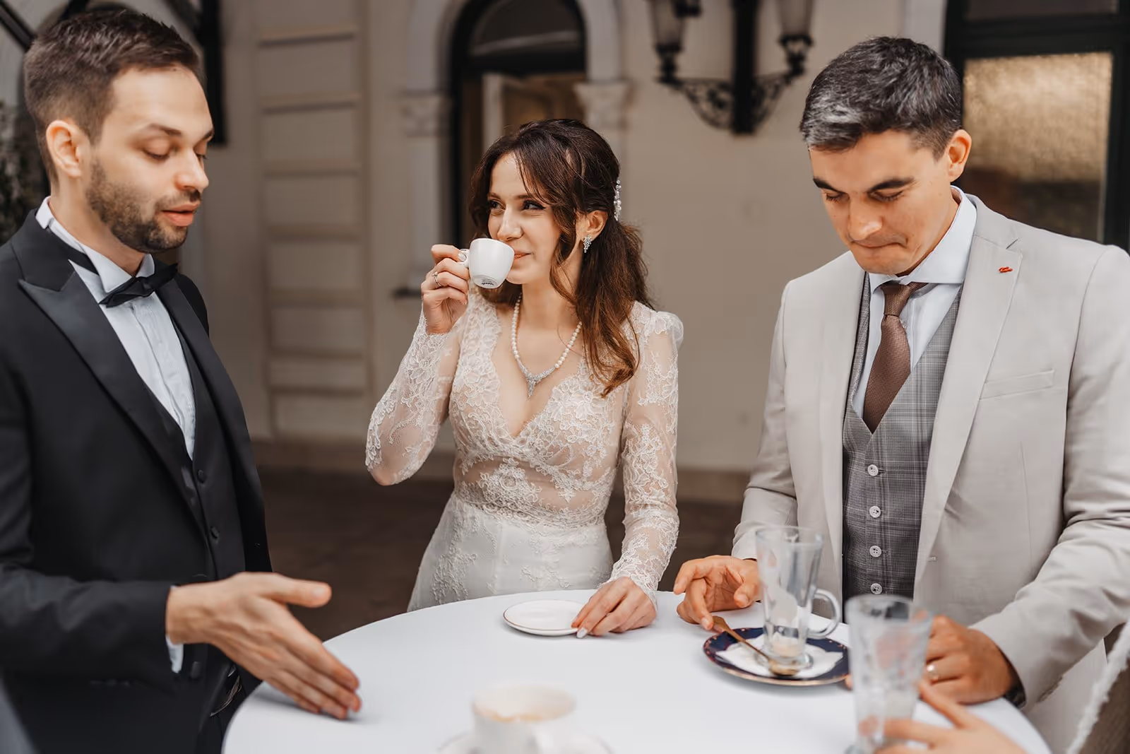 Bride in lace dress drinking from a small cup standing at a table with two men in formal suits.