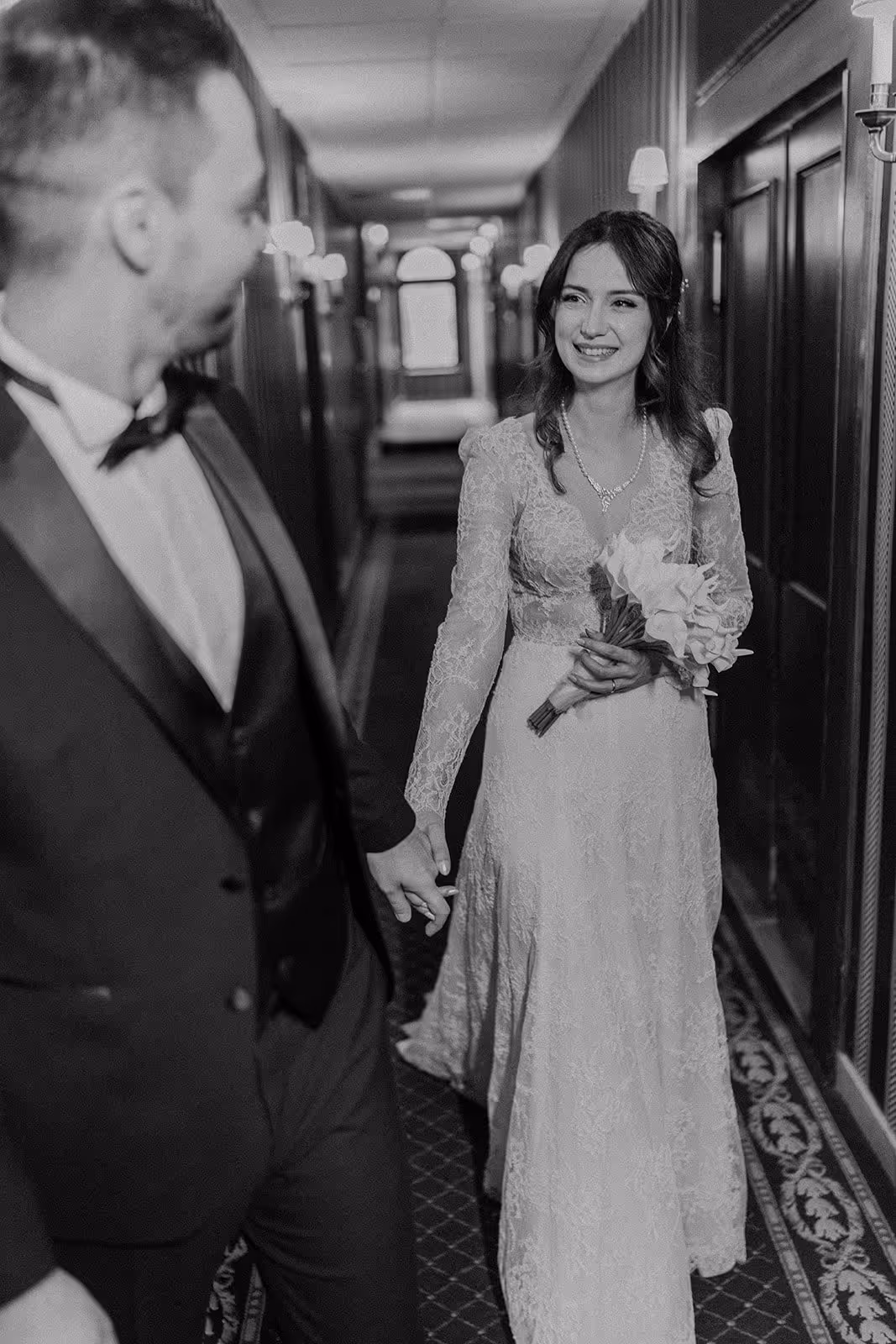 Bride and groom holding hands and smiling at each other in a hallway, bride holding a bouquet.