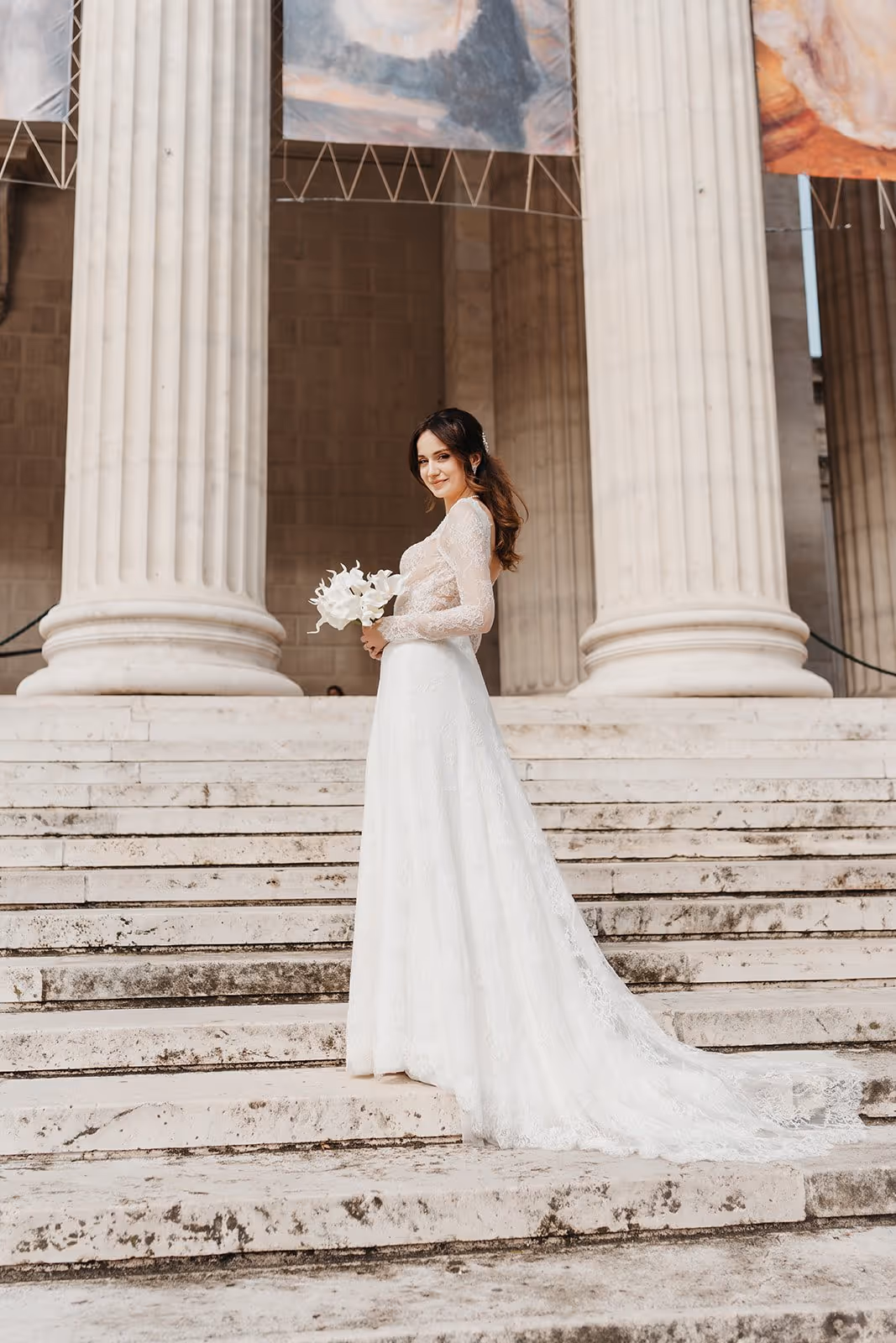 Bride in a long lace wedding dress holding a white bouquet, standing on stone steps with large columns in the background.