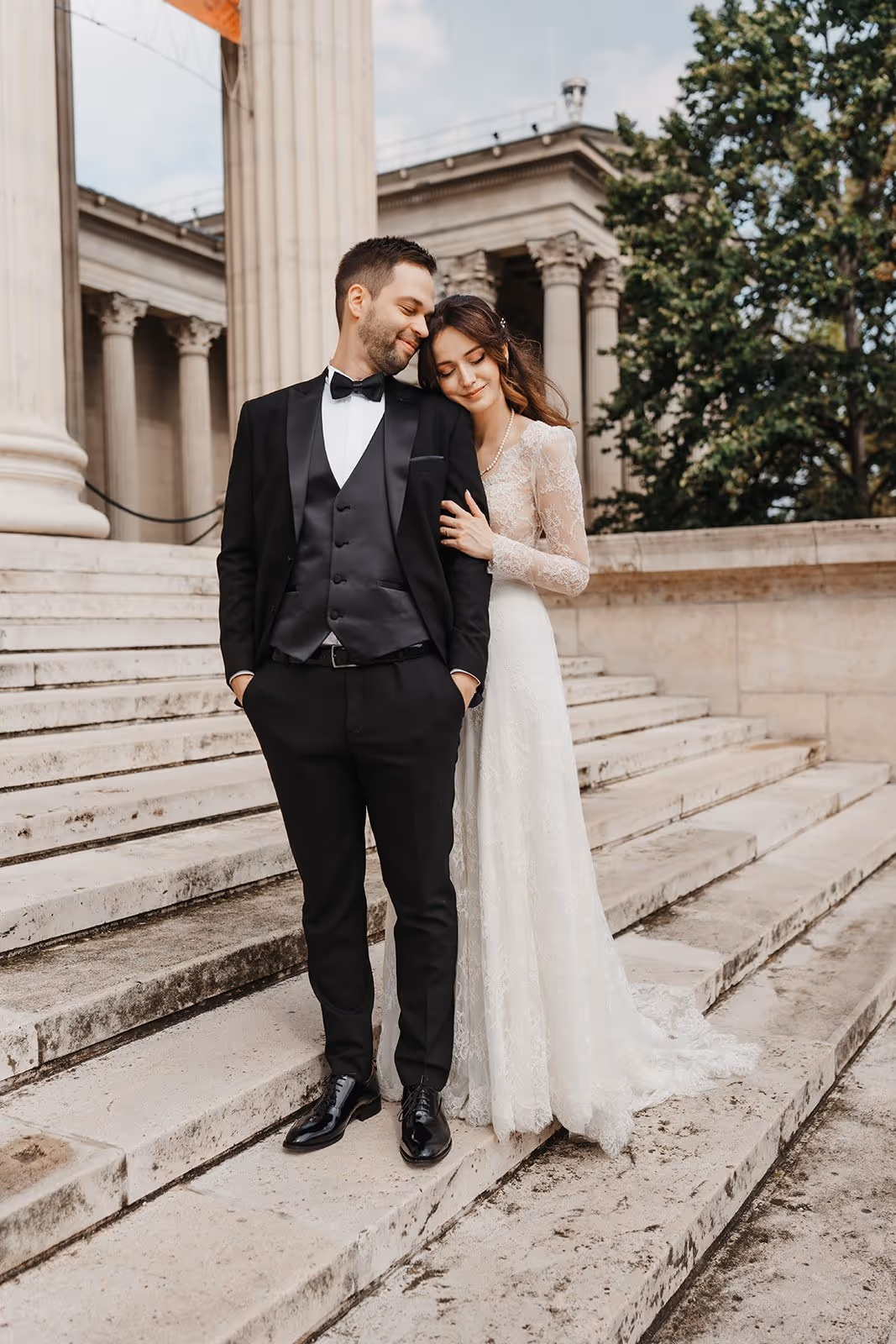 Bride in lace wedding dress leaning on groom in black tuxedo on stone steps outside a classical building.