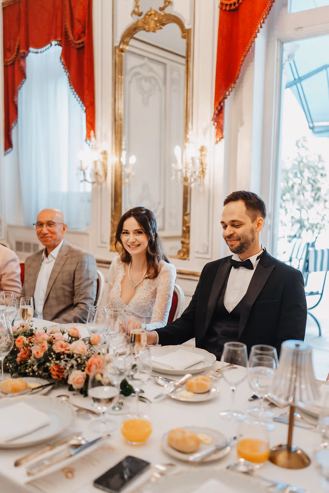 Bride in lace dress and groom in black tuxedo sitting at an elegantly set wedding reception table with floral centerpiece.