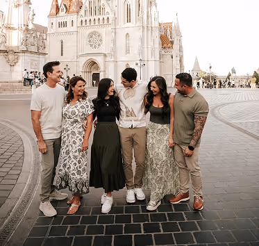 Five adults standing arm-in-arm on a paved square in front of a large historic building with ornate architecture.
